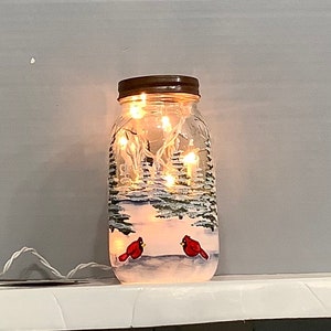 May include: A hand-painted mason jar with a winter scene, featuring two red cardinals and a snowy landscape. The jar is illuminated by fairy lights, creating a warm glow. The jar has a brown metal lid.