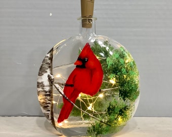 May include: A clear glass bottle with a cork top, containing a painted cardinal bird, a birch tree, and green foliage. The bottle is illuminated with white fairy lights.