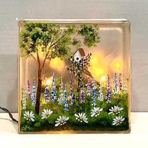 May include: A hand-painted glass block with a light-up feature, depicting a whimsical scene of a birdhouse nestled in a blooming garden with purple and blue lupine flowers and white daisies. The block is illuminated with warm white LED lights, creating a cozy and enchanting ambiance.