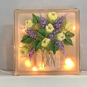 May include: A glass block with a painted image of a bouquet of yellow flowers and purple lilac in a white vase with a woven pattern. The block is illuminated with warm white LED lights.