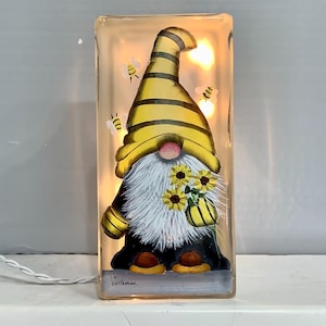 May include: A light-up glass block featuring a painted gnome with a yellow and black striped hat, holding sunflowers.  The gnome is surrounded by small bees.  The block is lit from within, creating a warm glow.  The signature "b. gentleman" is visible at the bottom of the block.