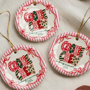 May include: Three round white ceramic ornaments with gold hanging strings. Each features a red and white striped border, a red ribbon illustration, and the text "COOKIE CREW" in patterned red and green letters, with "baking memories 2026" below.