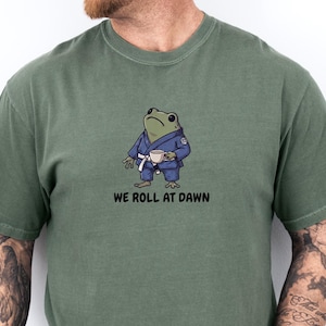 May include: An olive green short-sleeve t-shirt featuring a graphic of a green frog wearing a blue martial arts gi with a white belt and holding a small cup. Below the frog, the text reads WE ROLL AT DAWN in black, bold, sans-serif capital letters.