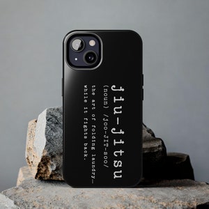 May include: Black phone case with the text "Jiu-jitsu" in white, along with a definition. The case is on a textured stone surface. The phone case has a camera and flash.