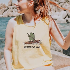 Pu&ograve; includere: Una canotta senza maniche di colore giallo chiaro con un'illustrazione grafica di una rana verde in piedi su una tavola da paddle mentre tiene un remo. Sotto l'illustrazione, il testo recita WE PADDLE AT DAWN in un semplice carattere nero.