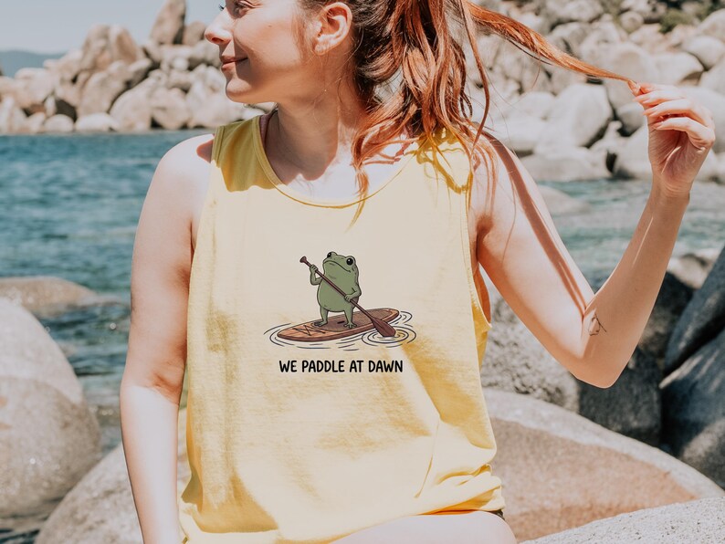 Pu&ograve; includere: Una canotta senza maniche di colore giallo chiaro con un'illustrazione grafica di una rana verde in piedi su una tavola da paddle mentre tiene un remo. Sotto l'illustrazione, il testo recita WE PADDLE AT DAWN in un semplice carattere nero.