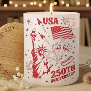 May include: A lit white candle with red graphics celebrating the USA's 250th Anniversary. The design includes the Statue of Liberty, an American flag, fireworks, and the word "USA". A wooden lid is next to the candle.