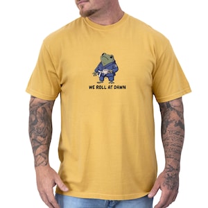 May include: A mustard yellow short-sleeve t-shirt featuring a graphic of a green frog wearing a blue martial arts gi with a white belt and holding a small white cup. Below the frog, the text reads WE ROLL AT DAWN in black capital letters.