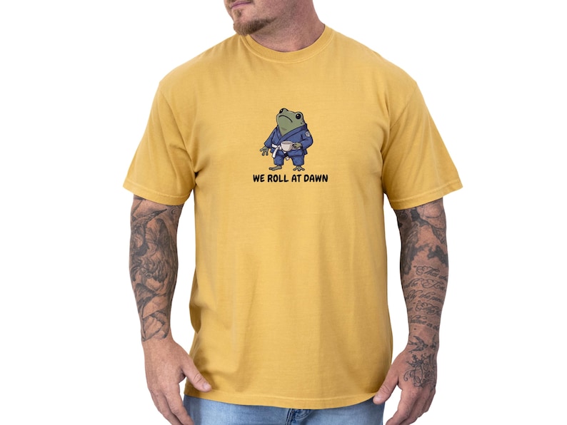 May include: A mustard yellow short-sleeve t-shirt featuring a graphic of a green frog wearing a blue martial arts gi with a white belt and holding a small white cup. Below the frog, the text reads WE ROLL AT DAWN in black capital letters.
