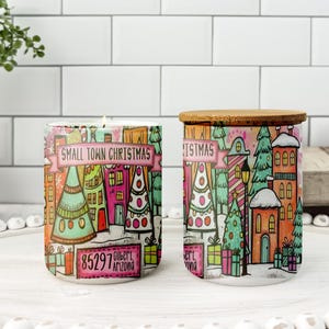 May include: Two ceramic candle holders with a Christmas village design. One has a lit candle. The design features colorful houses, trees, and the text "Small Town Christmas" and "85297 Gilbert, Arizona". The other has a wooden lid.