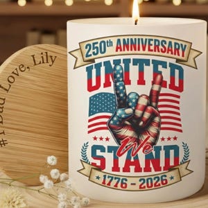 May include: A white candle with a flame, featuring a patriotic design for the 250th anniversary. The design includes a hand gesture in red, white, and blue, the American flag, and the text "United We Stand 1776-2026". A wooden coaster is next to the candle.