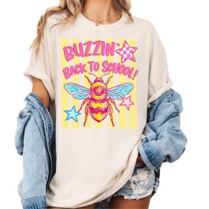 K&ouml;nnte beinhalten: Ein cremefarbenes T-Shirt mit einem Grafikdruck einer Biene in Rosa- und Gelbt&ouml;nen, blauen Fl&uuml;geln und gelben vertikalen Streifen. Der Text BUZZIN BACK TO SCHOOL ist in rosa Buchstaben gedruckt, erg&auml;nzt durch rosa und blaue Sterne.