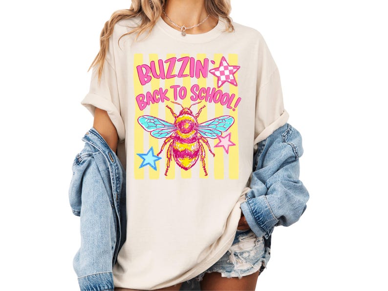 K&ouml;nnte beinhalten: Ein cremefarbenes T-Shirt mit einem Grafikdruck einer Biene in Rosa- und Gelbt&ouml;nen, blauen Fl&uuml;geln und gelben vertikalen Streifen. Der Text BUZZIN BACK TO SCHOOL ist in rosa Buchstaben gedruckt, erg&auml;nzt durch rosa und blaue Sterne.
