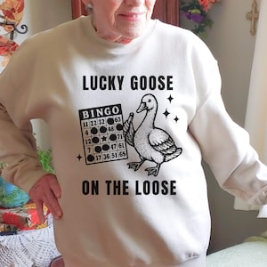 May include: A beige sweatshirt with the text "LUCKY GOOSE ON THE LOOSE" in black. The design includes a cartoon goose holding a bingo card. The bingo card has numbers and stars.