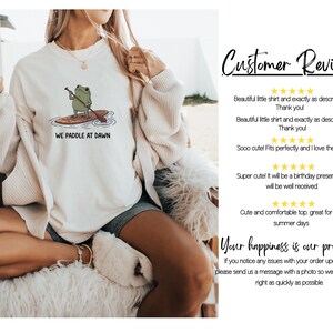 Pu&ograve; includere: Una felpa girocollo color crema con la grafica di una rana verde su una tavola da paddle e il testo "WE PADDLE AT DAWN". A destra, una sezione intitolata "Customer Reviews" mostra valutazioni a cinque stelle e commenti testuali.