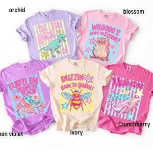 K&ouml;nnte beinhalten: F&uuml;nf kurz&auml;rmelige T-Shirts in den Farben Orchidee, Bl&uuml;te, Neonviolett, Elfenbein und Crunchberry. Jedes Shirt zeigt einen Grafikdruck mit Text: In the Door mit einem Dinosaurier, Whoooo's Ready for School mit einer Eule, Turtley Ready to Teach mit einer Schildkr&ouml;te, Buzzin Back to School mit einer Biene und Whale-come to Our Classroom mit einem Wal.