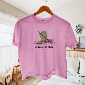 Pu&ograve; includere: Una t-shirt a maniche corte color rosa chiaro con una grafica raffigurante una rana verde in piedi su una tavola da paddle marrone nell'acqua. Il testo sotto la grafica recita WE PADDLE AT DAWN in lettere maiuscole nere su una gruccia.