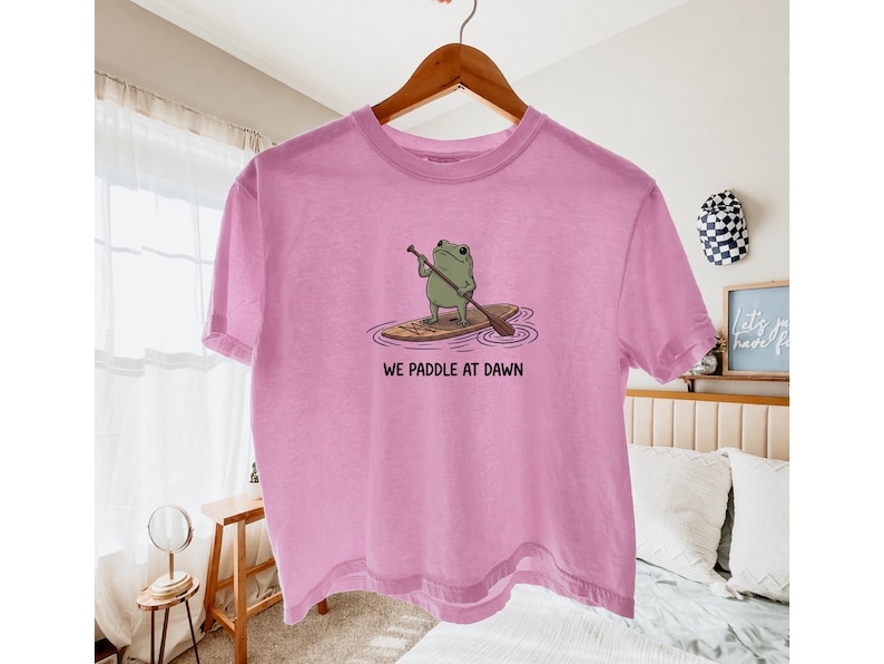 Pu&ograve; includere: Una t-shirt a maniche corte color rosa chiaro con una grafica raffigurante una rana verde in piedi su una tavola da paddle marrone nell'acqua. Il testo sotto la grafica recita WE PADDLE AT DAWN in lettere maiuscole nere su una gruccia.