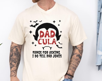 May include: Cream-colored t-shirt with a black and red graphic. The design features a vampire-like figure with the words "DAD CULA" and the phrase "FANGS FOR ASKING, I DO TELL DAD JOKES". Bats and stars complete the design.