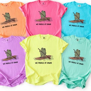 Pu&ograve; includere: Sei magliette a maniche corte con girocollo in vivaci colori neon tra cui arancione, rosa, blu, viola, giallo e verde menta. Ogni maglietta presenta una grafica di una rana verde su una tavola da paddle con il testo WE PADDLE AT DAWN stampato.