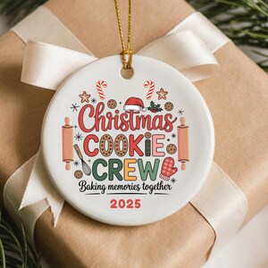 May include: A white ceramic Christmas ornament with a gold string, featuring the text "Christmas Cookie Crew" in red, brown, and green. The design includes candy canes, stars, and baking-themed graphics. The year 2025 is printed in red.