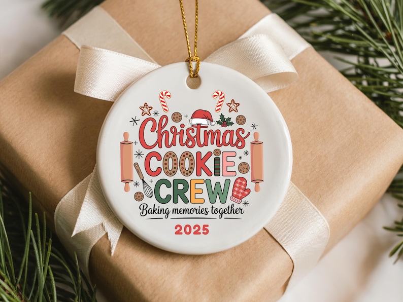 May include: A white ceramic Christmas ornament with a gold string, featuring the text "Christmas Cookie Crew" in red, brown, and green. The design includes candy canes, stars, and baking-themed graphics. The year 2025 is printed in red.