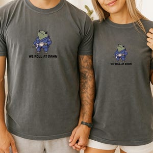 May include: A charcoal gray short-sleeve t-shirt featuring a graphic of a green frog wearing a blue martial arts gi and a white belt, holding a small white cup. Below the frog, the text reads WE ROLL AT DAWN in black capital letters.