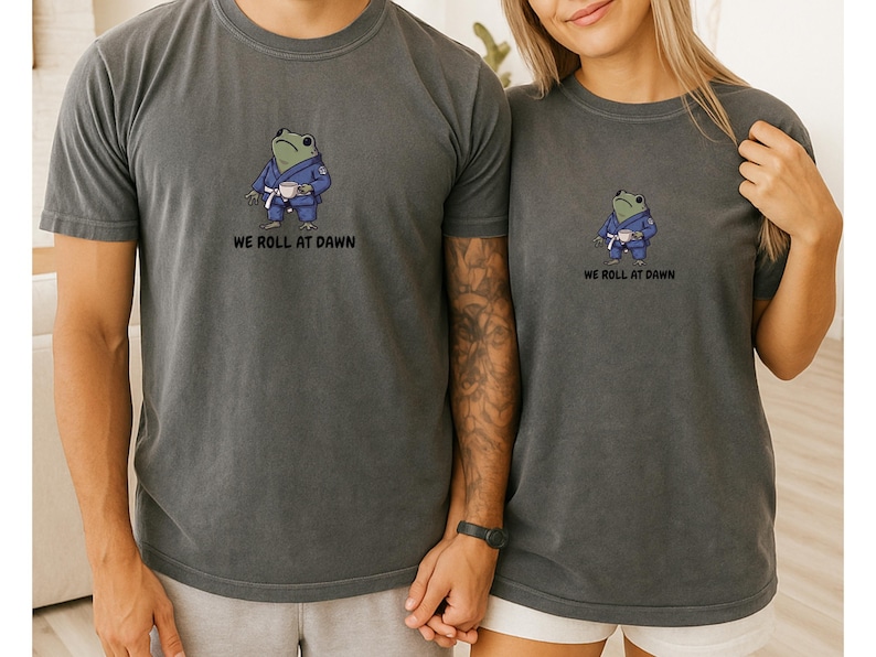 May include: A charcoal gray short-sleeve t-shirt featuring a graphic of a green frog wearing a blue martial arts gi and a white belt, holding a small white cup. Below the frog, the text reads WE ROLL AT DAWN in black capital letters.
