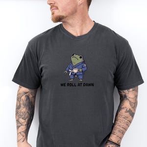 May include: A charcoal gray short-sleeve t-shirt featuring a graphic of a green frog wearing a blue martial arts gi with a white belt, holding a small white cup. Below the frog, the text reads WE ROLL AT DAWN in bold black capital letters.