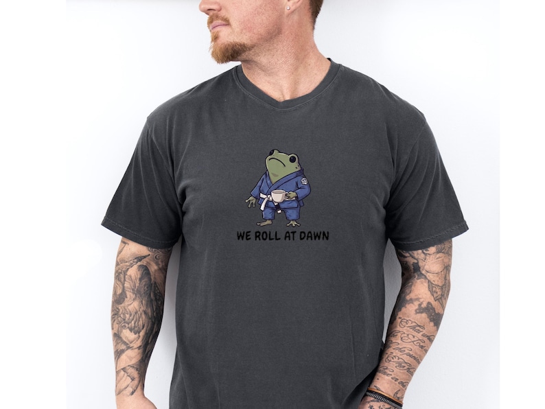 May include: A charcoal gray short-sleeve t-shirt featuring a graphic of a green frog wearing a blue martial arts gi with a white belt, holding a small white cup. Below the frog, the text reads WE ROLL AT DAWN in bold black capital letters.