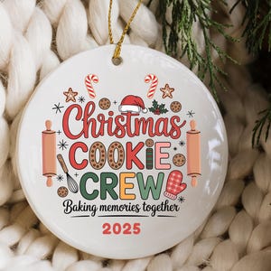 May include: A white ceramic Christmas ornament with a gold string. The ornament features the text "Christmas COOKIE CREW" in red, brown, green, and orange, with baking-themed illustrations. The text "Baking memories together 2025" is also included.