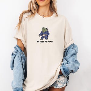 May include: A beige crew neck t-shirt featuring a graphic of a green frog wearing a blue martial arts gi and a white belt, holding a small white cup. Below the frog, the text reads WE ROLL AT DAWN in a bold, black, sans-serif font.