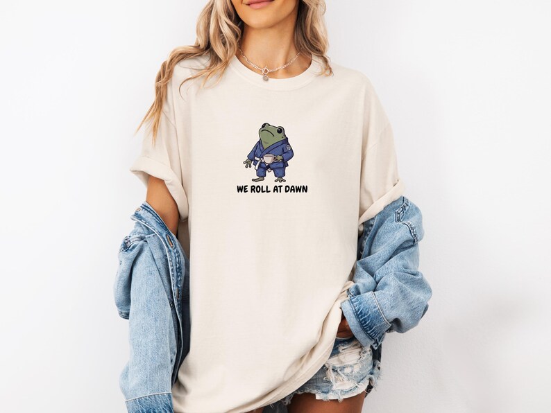 May include: A beige crew neck t-shirt featuring a graphic of a green frog wearing a blue martial arts gi and a white belt, holding a small white cup. Below the frog, the text reads WE ROLL AT DAWN in a bold, black, sans-serif font.