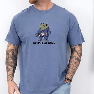 May include: A blue short-sleeved t-shirt featuring a graphic of a green frog wearing a blue martial arts gi with a white belt, holding a small white cup. Below the frog, the text reads WE ROLL AT DAWN in black, bold, sans-serif capital letters.