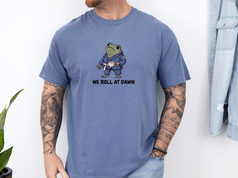 May include: A blue short-sleeved t-shirt featuring a graphic of a green frog wearing a blue martial arts gi with a white belt, holding a small white cup. Below the frog, the text reads WE ROLL AT DAWN in black, bold, sans-serif capital letters.