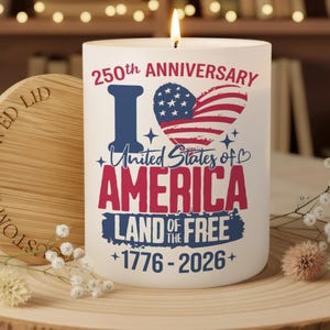 May include: A white candle with a lit flame, featuring patriotic text and graphics. The design includes the words "250th Anniversary," "I Love America," and "Land of the Free," with the years 1776-2026. A wooden lid is next to the candle.