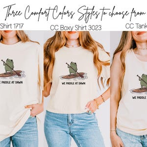 Pu&ograve; includere: Tre magliette color crema con la grafica di una rana verde su un paddleboard e il testo "WE PADDLE AT DAWN". Le opzioni includono una maglietta standard etichettata CC Shirt 1717, una maglietta boxy CC Boxy Shirt 3023 e una canotta CC Tank 9360.