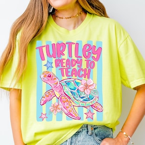 K&ouml;nnte beinhalten: Ein neongelbes Kurzarm-T-Shirt mit einer Grafik einer rosa Meeresschildkr&ouml;te mit buntem Panzer und Hibiskusbl&uuml;ten. Der Text "TURTLEY READY TO TEACH" steht in rosa Buchstaben vor einem Hintergrund aus hellblauen vertikalen Streifen.