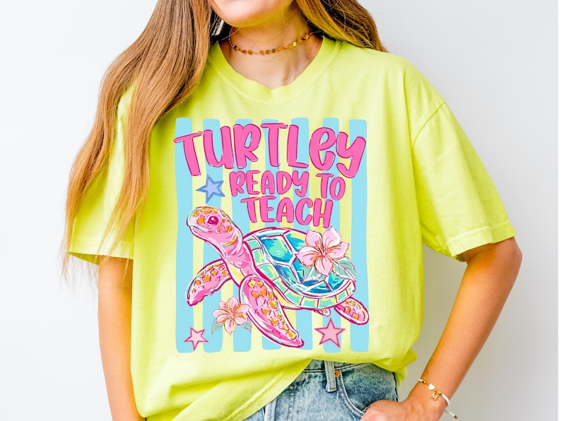 K&ouml;nnte beinhalten: Ein neongelbes Kurzarm-T-Shirt mit einer Grafik einer rosa Meeresschildkr&ouml;te mit buntem Panzer und Hibiskusbl&uuml;ten. Der Text "TURTLEY READY TO TEACH" steht in rosa Buchstaben vor einem Hintergrund aus hellblauen vertikalen Streifen.