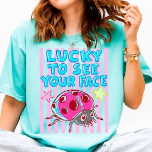K&ouml;nnte beinhalten: Ein t&uuml;rkisfarbenes T-Shirt mit einem Grafikdruck, der den Text "LUCKY TO SEE YOUR FACE" in blauen Buchstaben zeigt. Das Design enth&auml;lt einen rosa-roten Marienk&auml;fer, einen rosa Stern und einen gelben Stern auf vertikalen rosa Streifen.