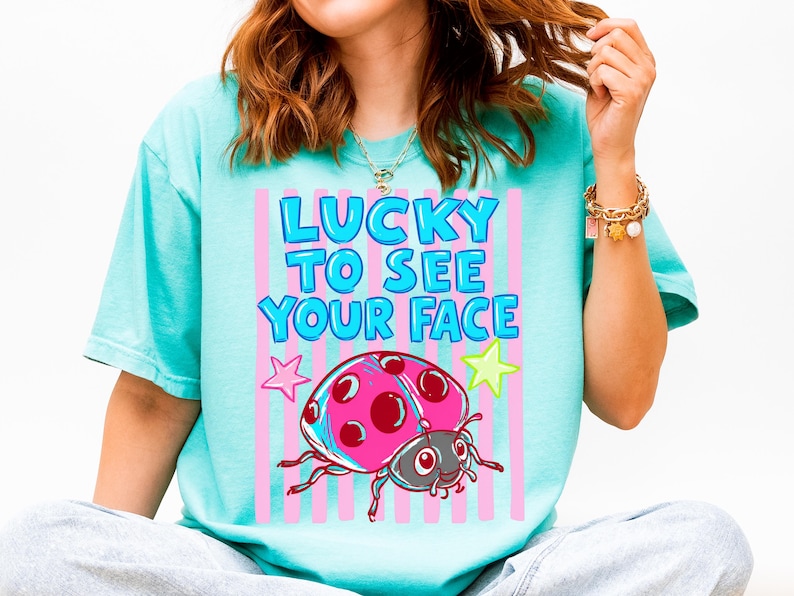 K&ouml;nnte beinhalten: Ein t&uuml;rkisfarbenes T-Shirt mit einem Grafikdruck, der den Text "LUCKY TO SEE YOUR FACE" in blauen Buchstaben zeigt. Das Design enth&auml;lt einen rosa-roten Marienk&auml;fer, einen rosa Stern und einen gelben Stern auf vertikalen rosa Streifen.