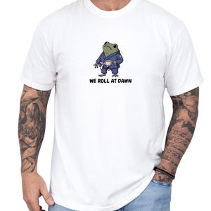 May include: A white t-shirt featuring a graphic of a green frog wearing a blue martial arts gi with a white belt, holding a small cup. Below the frog, the text reads WE ROLL AT DAWN in black capital letters. The shirt has short sleeves and a crew neck.