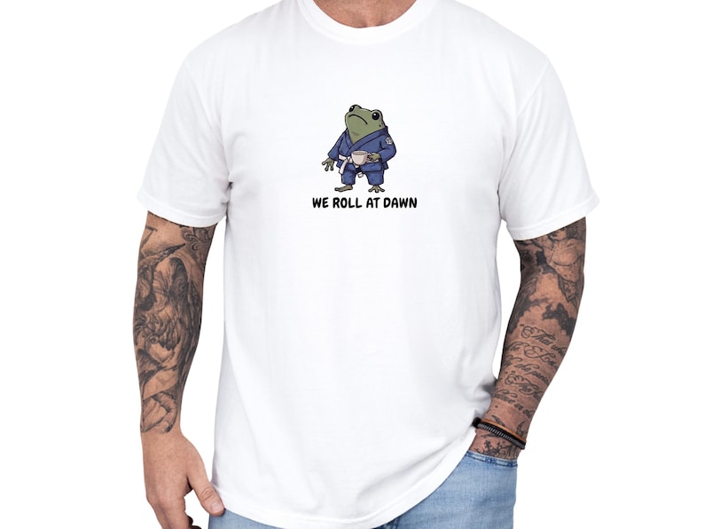 May include: A white t-shirt featuring a graphic of a green frog wearing a blue martial arts gi with a white belt, holding a small cup. Below the frog, the text reads WE ROLL AT DAWN in black capital letters. The shirt has short sleeves and a crew neck.