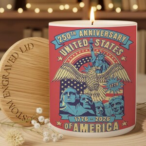 May include: A lit candle in a white cylindrical holder with a red graphic commemorating the 250th anniversary of the United States. The design includes an eagle, the Statue of Liberty, and the text "1776-2026 OF AMERICA". A wooden lid is next to the candle.