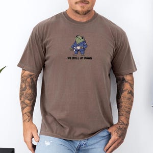 May include: A brown short-sleeve t-shirt featuring a graphic of a green frog wearing a blue martial arts gi and a white belt while holding a small white cup. Below the frog, the text reads WE ROLL AT DAWN in black, bold, sans-serif capital letters.