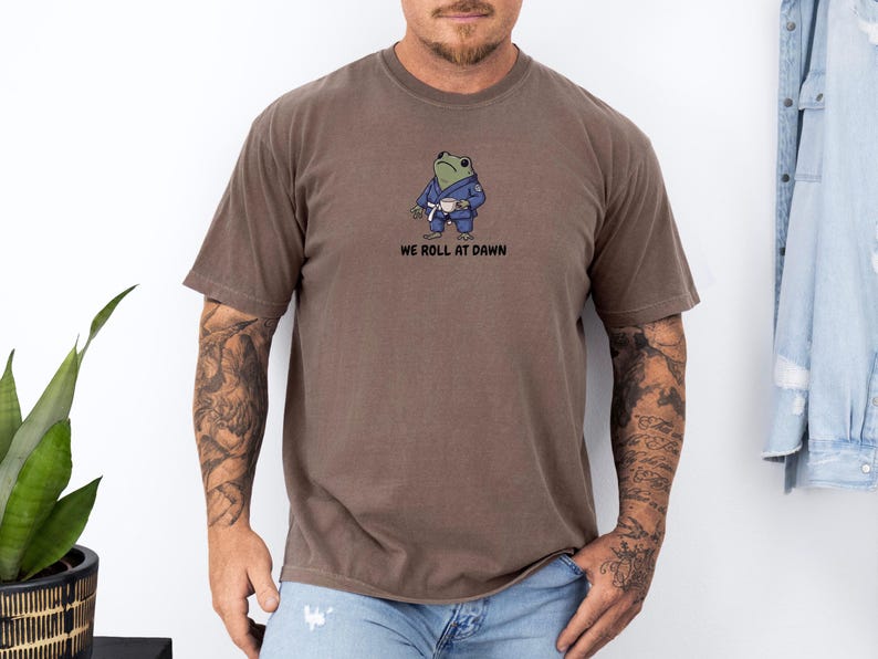May include: A brown short-sleeve t-shirt featuring a graphic of a green frog wearing a blue martial arts gi and a white belt while holding a small white cup. Below the frog, the text reads WE ROLL AT DAWN in black, bold, sans-serif capital letters.