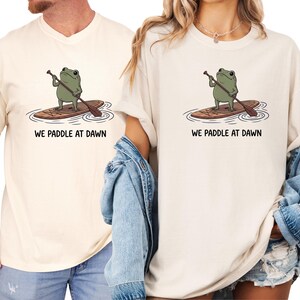 Pu&ograve; includere: Una t-shirt color crema con la grafica di una rana verde in piedi su una tavola da paddle marrone con un remo. Sotto l'illustrazione, il testo WE PADDLE AT DAWN &egrave; stampato in lettere maiuscole nere al centro della parte anteriore.