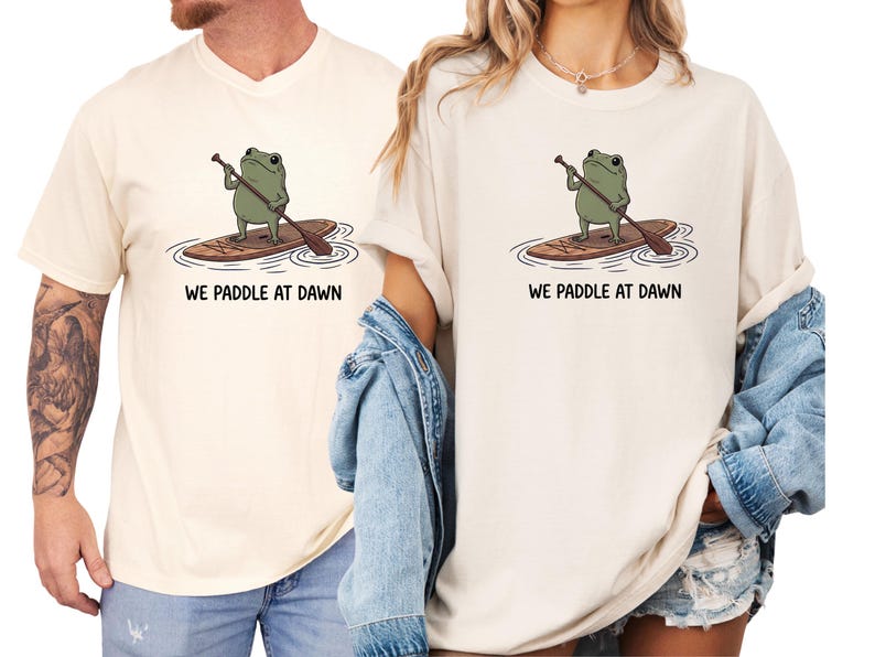 Pu&ograve; includere: Una t-shirt color crema con la grafica di una rana verde in piedi su una tavola da paddle marrone con un remo. Sotto l'illustrazione, il testo WE PADDLE AT DAWN &egrave; stampato in lettere maiuscole nere al centro della parte anteriore.