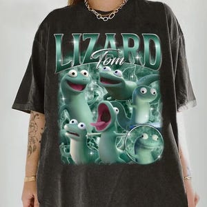 May include: Dark gray t-shirt featuring a graphic of several green cartoon lizards with the text "Lizard Tom" in a teal, retro font. The lizards have various expressions, and the design has a vintage, distressed look.