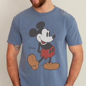 May include: Blue t-shirt featuring a classic Mickey Mouse graphic. The cartoon character is depicted in black, white, red, and tan, with the Disney logo printed below. The shirt has short sleeves and a crew neck.